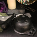 6Witch3 Cast Iron Cauldron with Pestle - Shown with the cauldron flipped upside down so you can see its three feet. The pestle rests nearby. In the background are a selenite incense burner, a candle, a purple rose, an abalone shell, and a small pair of metal tongs.