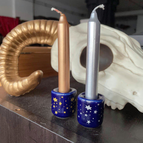 6Witch3 Metallic Chime Candles - a pair of cobalt blue small candle holders rest on a wood cabinet next to a not entirely realistic resin cow skull. One holder has gold stars and the other has silver stars, each holder is holding a chime candle that matches its stars.