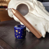 6Witch3 Metallic Chime Candles - a cobalt blue porcelain candle holder sits next to a somewhat cartoonish cow skull model, the holder has gold stars and a gold candle leans against it. The gold is a warm coppery color with more of an orange tone than a yellow.