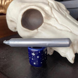 6Witch3 Metallic Chime Candles - a cobalt blue porcelain candle holder sits next to a somewhat cartoonish cow skull model. The holder has silver stars and a silver candle lays atop the holder. The silver of the candle is a lovely pearlescent silver, not too warm or cool in tone.