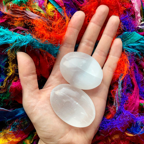 6Witch3 Selenite Palm Stones - two milky semi-transparent stones rest in the palm of an open hand held over a puddle of frayed, brightly colored silk strips.