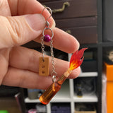 A hand holding an earring with a tiny bottle with a tiny flame shooting out of it, and a tiny brick. Each charm is on a thin steel chain and each chain is on a jump ring on a French earring hook.