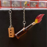 A close image of one tiny brick on a chain with an earring hook, and one tiny bottle with a tiny flame coming out of it.