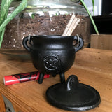 6Witch3 black iron cauldron with a pentacle design on the side. It's shown next to a potted aloe, its lid is next to it and it has a bundle of herbs in it with a red lighter nearby. 