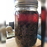 6Witch3 Agua Florida Florida Water - a glass quart jar half full of botanicals and a dark reddish brown liquid stands on a wooden box by a window so that the light shines through the liquid inside