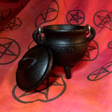6Witch3 Mini Cast Iron Painted Cauldron - a small round black cauldron stands on a red background spangled with lack pentacles. The cauldron's lid is off to the cauldron's side, leaning against it at a jaunty angle. 