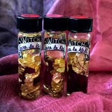 6Witch3 Scent Oil Sara La Kali - three 2 dram bottles of oil stand on a purple and pink pile of silk scarf. The oil bottles have a bit of rose petal and 24k gold leaf in the bottles along with the slightly golden oil. 