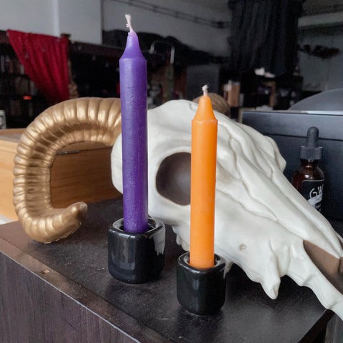 6Witch3 Plain Black Porcelain Chime Holder - a large purple chime candle in a shiny black porcelain holder sits with a small orange chime candle in a small shiny cube-shaped black holder. The candles are sitting next to a cartoonish resin replica of a cow skull and all are resting on a dark brown wood cabinet.