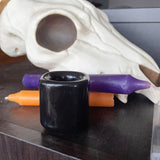6Witch3 Plain Black Porcelain Chime Holder - a large purple candle and a small orange candle lay in an overlapping pile next to a roughly attempted plastic cow skull. In front of the candles and skull is a shiny black square glass chime holder without a candle in it. This tableau is displayed on a dark wood cabinet.