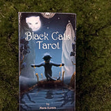 6Witch3 Black Cats Tarot - The front of the box shows a black cat standing on a dock with its back to the water, arms outstretched, looking up at a moon whose face is that of a white cat. The black cat is wearing gold bracelets on each wrist and one ring around the tip of its tail.