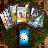 6Witch3 Black Cats Tarot - A spread of cards next to a facedown stack of cards, both arrayed on a round patch of green moss resting on a brown wooden background.