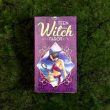 6Witch3 Teen Witch Tarot - The purple card box lays on a circle of green moss. The box images shows a young witch in a traditional large purple witch hat and a maroon wrap halter top peering into a hand mirror. A book is open on their lap.