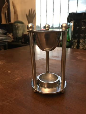 6Witch3 Hourglass Oil Burner - a silver oil diffuser sits on a wooden table, three pencil-thin columns with tiny pea-sized balls on top support a silver cup with a space at the base for a single tea light.